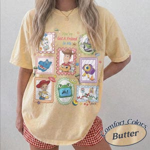 May include: A pale yellow short-sleeve t-shirt featuring a graphic print of various animated characters in postage stamp-style frames. The text "You've Got A Friend In Me" is printed in the center. The design includes colorful illustrations.