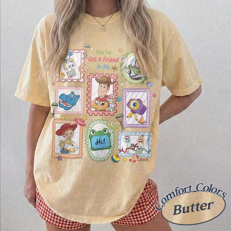 May include: A pale yellow short-sleeve t-shirt featuring a graphic print of various animated characters in postage stamp-style frames. The text "You've Got A Friend In Me" is printed in the center. The design includes colorful illustrations.