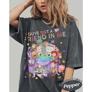 May include: A charcoal gray short-sleeve t-shirt featuring a colorful graphic print of various animated toy characters and the text "YOU'VE GOT A FRIEND IN ME" at the top. The design includes a purple bag, stars, and blocks on a vintage-wash fabric.