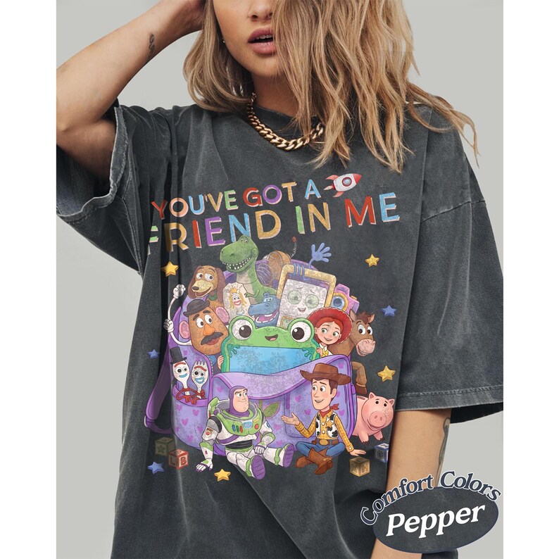 May include: A charcoal gray short-sleeve t-shirt featuring a colorful graphic print of various animated toy characters and the text "YOU'VE GOT A FRIEND IN ME" at the top. The design includes a purple bag, stars, and blocks on a vintage-wash fabric.