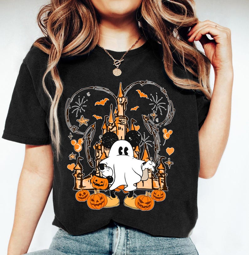 May include: Black t-shirt with a Halloween design. The graphic features a castle, a ghost, pumpkins, and Mickey Mouse ears. The design uses orange, white, and black, with fireworks and bats. This casual t-shirt is suitable for Halloween.