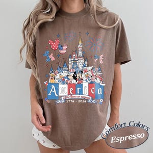 May include: A brown t-shirt featuring a graphic of a castle, fireworks, and cartoon characters wearing patriotic red, white, and blue clothing. The text reads America, 250 Years of Freedom, and 1776-2026. The shirt is labeled as Comfort Colors Espresso.