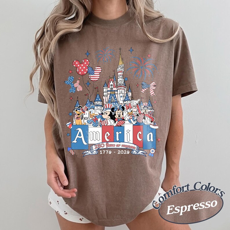 May include: A brown t-shirt featuring a graphic of a castle, fireworks, and cartoon characters wearing patriotic red, white, and blue clothing. The text reads America, 250 Years of Freedom, and 1776-2026. The shirt is labeled as Comfort Colors Espresso.