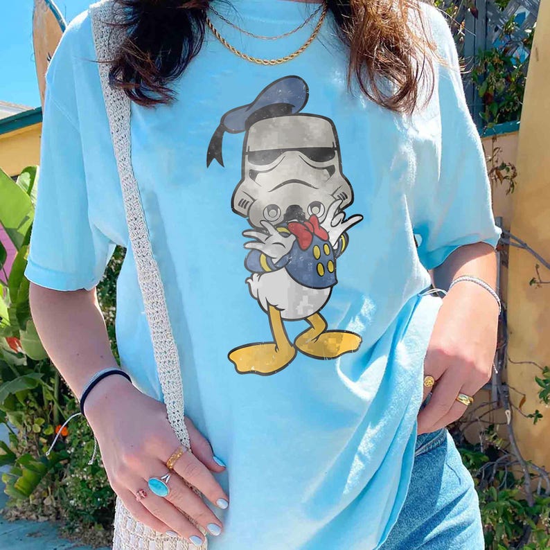 May include: Light blue t-shirt featuring a graphic of Donald Duck dressed as a Stormtrooper. The cartoon character wears a Stormtrooper helmet and a blue sailor suit with a red bow tie. The shirt has short sleeves and a crew neck.