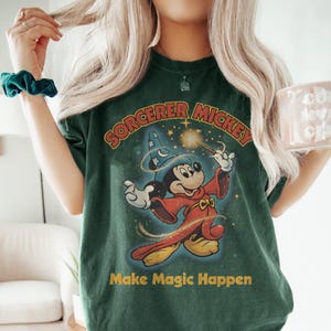 Peut inclure: T-shirt vert fonc&eacute; avec Mickey Mouse en sorcier tenant une baguette magique. Le design comprend le texte "SORCERER MICKEY" et "Make Magic Happen" en jaune. Le t-shirt a un aspect vintage et vieilli.