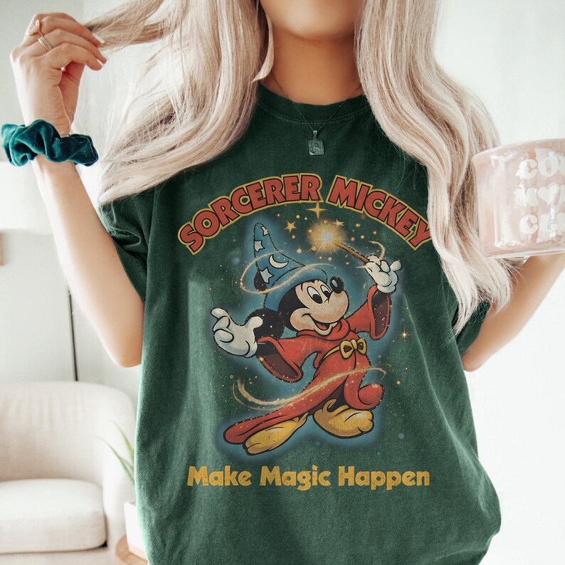 Peut inclure: T-shirt vert fonc&eacute; avec Mickey Mouse en sorcier tenant une baguette magique. Le design comprend le texte "SORCERER MICKEY" et "Make Magic Happen" en jaune. Le t-shirt a un aspect vintage et vieilli.
