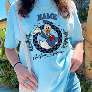 K&ouml;nnte beinhalten: Hellblaues T-Shirt mit einem Cartoon-Design von Donald Duck in Marineblau und Rot. Das Shirt hat das Wort "NAME" &uuml;ber dem Bild und "Custom Name" darunter. Das Design befindet sich in einem karierten Kreis.