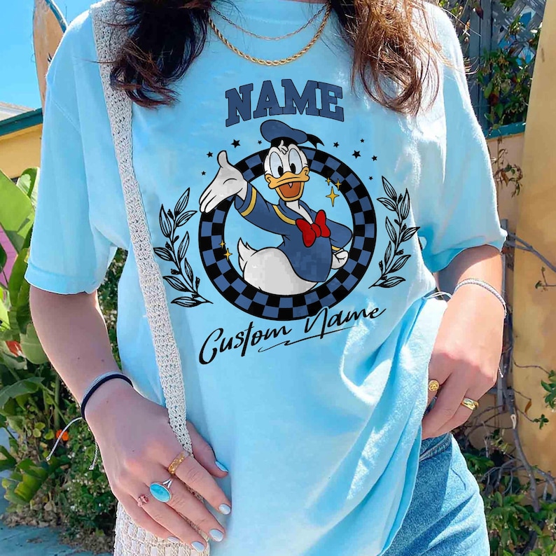 K&ouml;nnte beinhalten: Hellblaues T-Shirt mit einem Cartoon-Design von Donald Duck in Marineblau und Rot. Das Shirt hat das Wort "NAME" &uuml;ber dem Bild und "Custom Name" darunter. Das Design befindet sich in einem karierten Kreis.