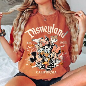 K&ouml;nnte beinhalten: Orangefarbenes T-Shirt mit einem Motiv von Mickey Mouse, Donald Duck, Goofy und Pluto, die als Skelette verkleidet sind. Der Text "Disneyland Est 1955 California" ist auf dem Shirt aufgedruckt.