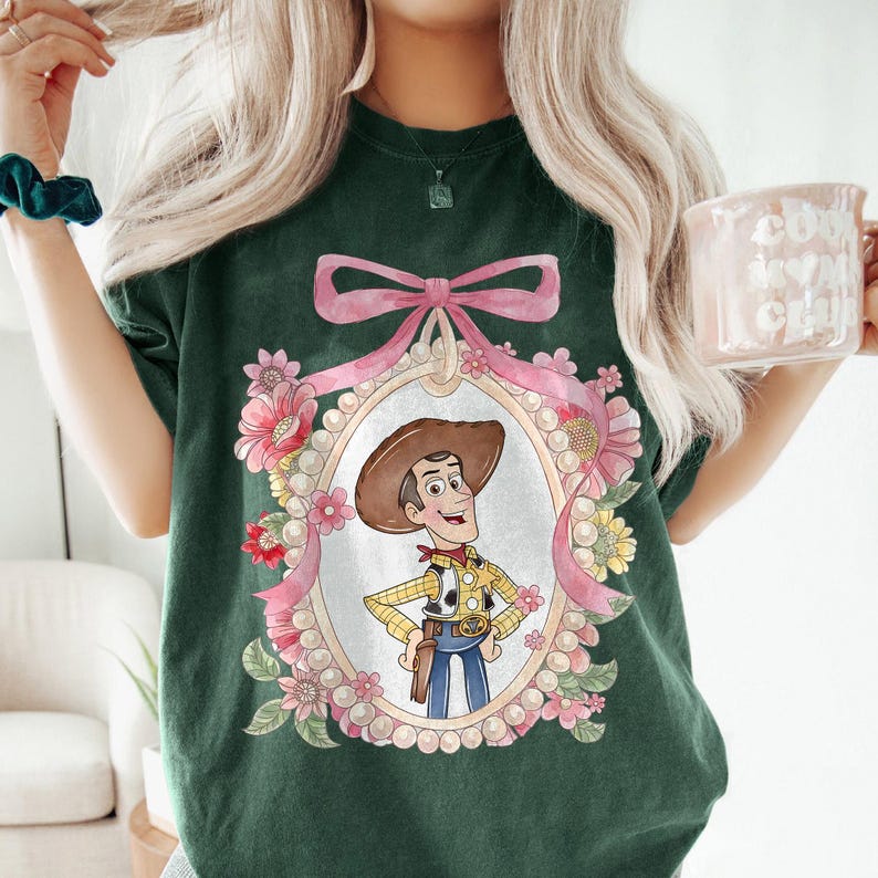 May include: Dark green t-shirt featuring a cartoon image of Woody from Toy Story. The image is framed with a pink bow and floral accents. Woody is wearing a cowboy hat, yellow shirt, blue jeans, and a holster. The shirt is a comfortable, casual style.