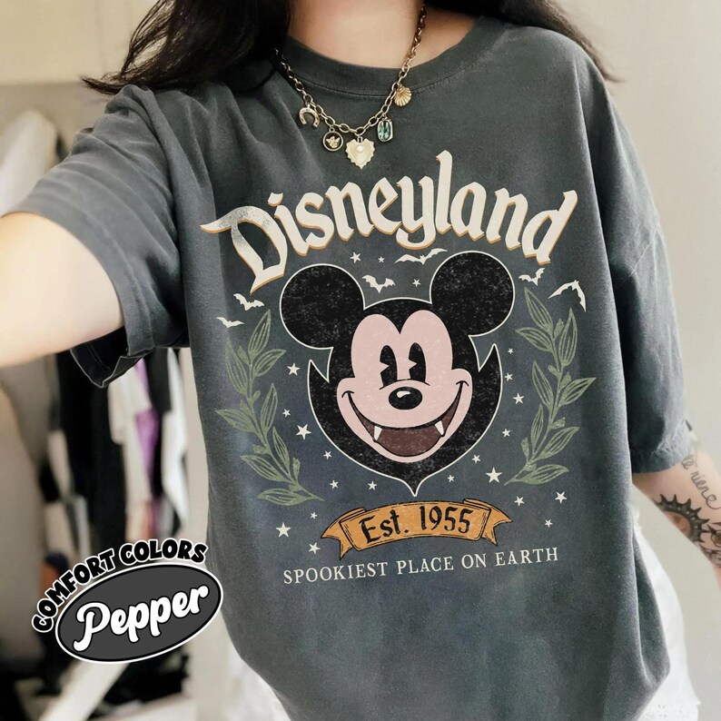 May include: A dark gray t-shirt with a Disneyland graphic featuring Mickey Mouse with fangs. The design includes the text "Disneyland", "Est. 1955", and "Spookiest Place on Earth". The shirt is from the Comfort Colors brand.