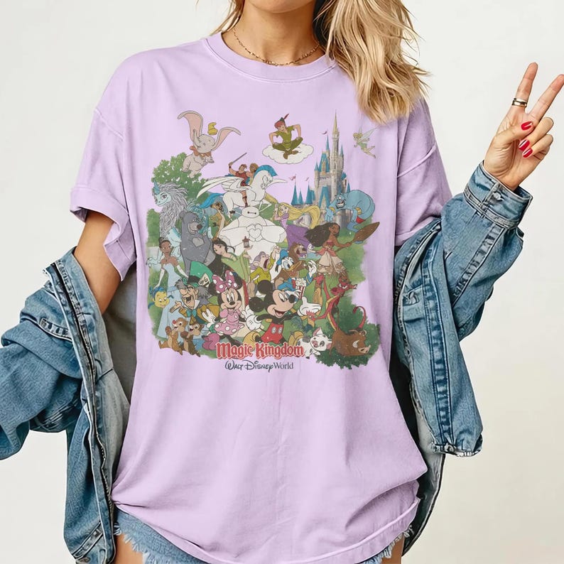 K&ouml;nnte beinhalten: Ein lila T-Shirt mit einer farbenfrohen Grafik verschiedener Disney-Charaktere, darunter Mickey und Minnie Mouse, Peter Pan und Dumbo. Der Text "Magic Kingdom" und "Walt Disney World" ist darunter gedruckt.