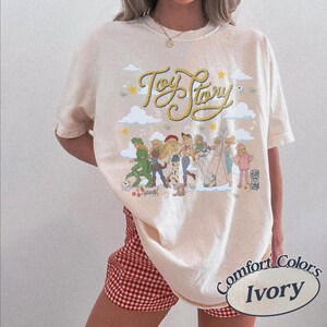 May include: An ivory short-sleeve t-shirt featuring a graphic print with the text Toy Story in yellow rope-style lettering. The design includes illustrations of various characters, clouds, and stars. The shirt has a crew neckline and a relaxed fit.