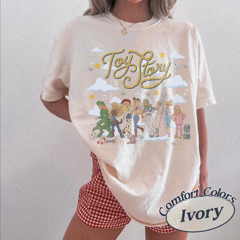May include: An ivory short-sleeve t-shirt featuring a graphic print with the text Toy Story in yellow rope-style lettering. The design includes illustrations of various characters, clouds, and stars. The shirt has a crew neckline and a relaxed fit.
