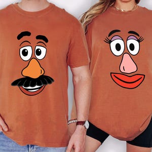 May include: Two rust-coloured t-shirts featuring cartoon faces. One shirt has a moustache, orange nose, and a smiling mouth. The other shirt has eyelashes, a pink nose, and a red smiling mouth. The shirts are likely part of a matching set.