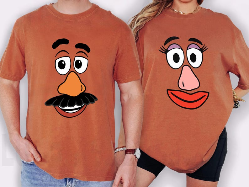 May include: Two rust-coloured t-shirts featuring cartoon faces. One shirt has a moustache, orange nose, and a smiling mouth. The other shirt has eyelashes, a pink nose, and a red smiling mouth. The shirts are likely part of a matching set.