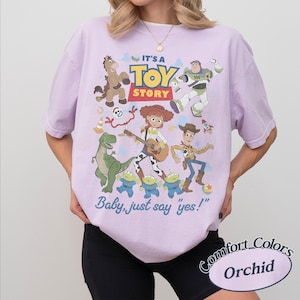 May include: A light purple t-shirt featuring a graphic print of Toy Story characters including Woody, Buzz Lightyear, Jessie, Bullseye, Rex, Forky, and the aliens. The text reads IT'S A TOY STORY and Baby, just say yes! in blue and yellow lettering.