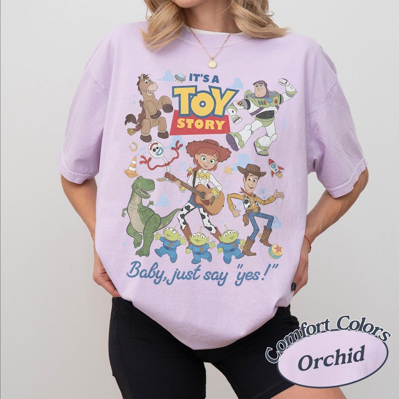May include: A light purple t-shirt featuring a graphic print of Toy Story characters including Woody, Buzz Lightyear, Jessie, Bullseye, Rex, Forky, and the aliens. The text reads IT'S A TOY STORY and Baby, just say yes! in blue and yellow lettering.