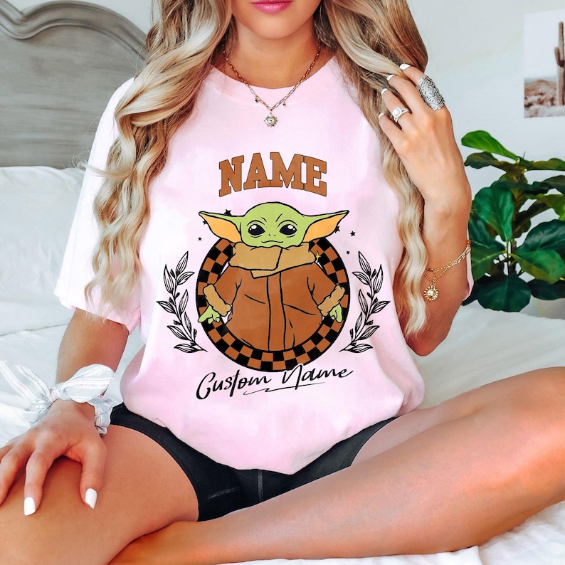 K&ouml;nnte beinhalten: Hellrosa T-Shirt mit einem gr&uuml;nen Alien-Charakter in einem braunen Mantel, umgeben von einem schwarz-braunen karierten Kreis und Lorbeerbl&auml;ttern. Das Wort "NAME" steht &uuml;ber der Figur und "Custom Name" darunter.