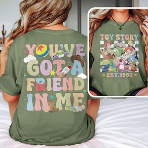 May include: Green t-shirt with a retro-style graphic that says "You've Got a Friend in Me" with a cartoon image of Woody's hat, a rocket, a horseshoe, and a planet. The back of the shirt has a checkered pattern with a group of Toy Story characters and the text "Toy Story Est. 1995".