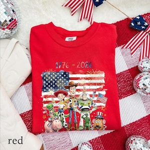 May include: A red crewneck t-shirt featuring a graphic print of various animated toy characters standing in front of an American flag with fireworks. The text 1776 - 2026 is printed above the characters in blue and red colors.
