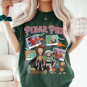May include: Dark green t-shirt with the words "PIXAR PIER" in red and white lettering. The shirt features images of various Pixar characters, including Woody, Buzz Lightyear, and others, with a spiderweb design.