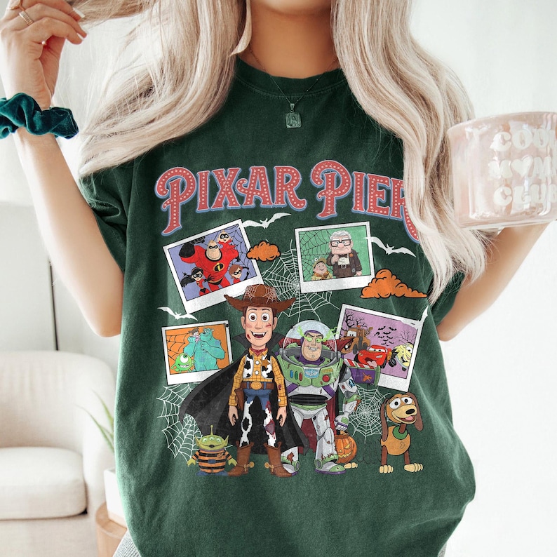 May include: Dark green t-shirt with the words "PIXAR PIER" in red and white lettering. The shirt features images of various Pixar characters, including Woody, Buzz Lightyear, and others, with a spiderweb design.