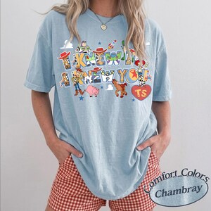 May include: A light blue short-sleeve t-shirt featuring a graphic print with the text I KNEW I KNEW YOU. The design includes colorful illustrations of various animated toy characters, a rocket, stars, and a red heart shape containing the letters TS.
