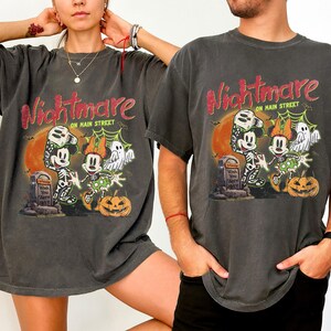 May include: Dark gray t-shirts with a Halloween-themed graphic. The design features Mickey and Minnie Mouse in skeleton costumes, a ghost, and a jack-o'-lantern, with the words "Nightmare on Main Street".