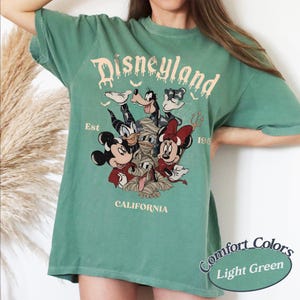 May include: A light green short-sleeve t-shirt featuring a graphic print of Disney characters in Halloween costumes. The text reads Disneyland Est 1955 California. The characters include Mickey, Minnie, Donald, Goofy, and Pluto as a mummy.
