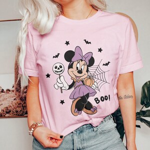 May include: A light pink t-shirt featuring a Halloween-themed Minnie Mouse graphic. Minnie is dressed in purple and holding a skull lollipop and a pumpkin. The word "BOO!" is printed on the shirt, along with bats and stars.