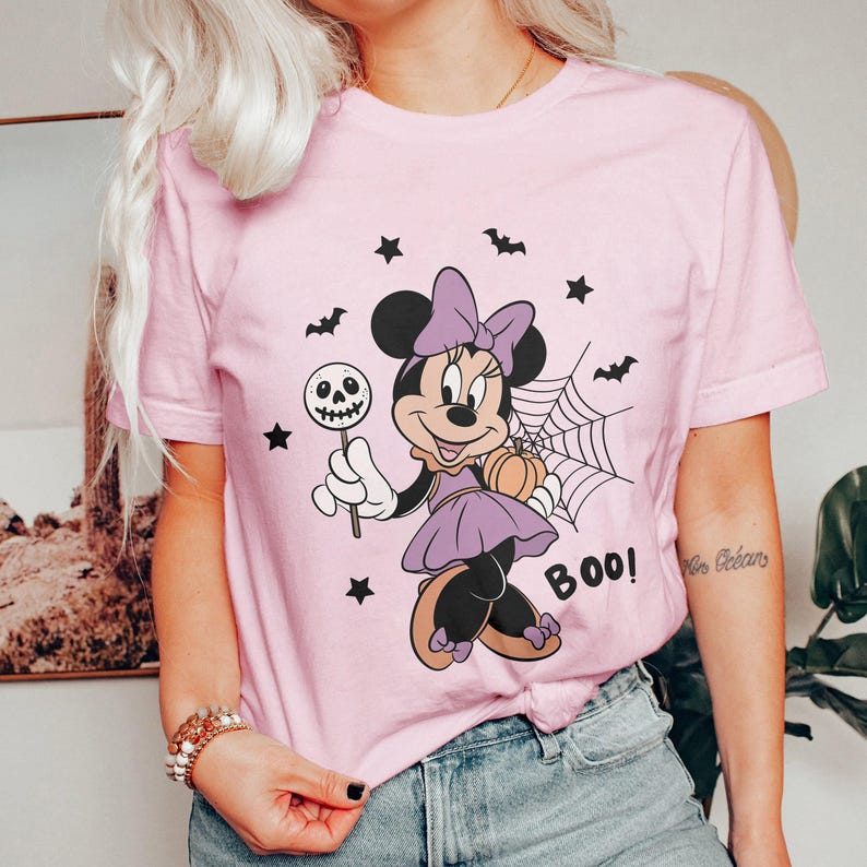 May include: A light pink t-shirt featuring a Halloween-themed Minnie Mouse graphic. Minnie is dressed in purple and holding a skull lollipop and a pumpkin. The word "BOO!" is printed on the shirt, along with bats and stars.