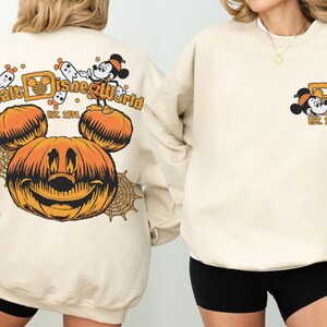 May include: Beige sweatshirt featuring a Halloween-themed Walt Disney World design. The back displays a large orange Mickey Mouse pumpkin, ghosts, and the text "Walt Disney World Est. 1971." The front has a smaller Mickey Mouse graphic. A cozy, festive apparel item.