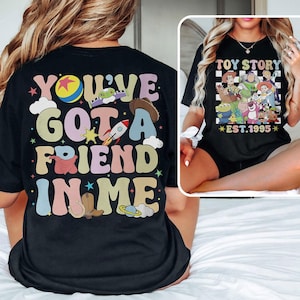 May include: Black t-shirt with a colorful retro graphic that says "You've Got a Friend in Me" with a cartoon rocket, cowboy hat, and stars. The back of the shirt has a checkered pattern with the text "Toy Story Est. 1995" and cartoon characters from the movie.
