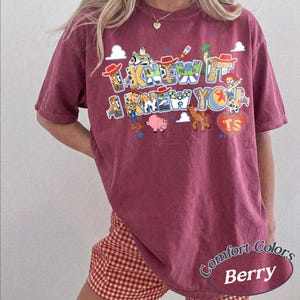May include: A berry-colored short-sleeve t-shirt featuring a graphic print of Toy Story characters including Woody, Buzz Lightyear, Jessie, Bullseye, and Hamm. The text reads I KNEW IT I KNEW YOU with a heart icon containing the letters TS.