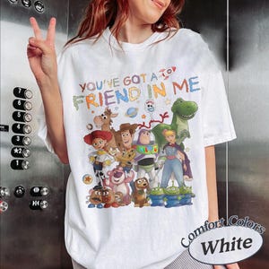 May include: A white short-sleeved t-shirt featuring a colorful graphic print of various animated characters from the Toy Story franchise. The text "YOU'VE GOT A FRIEND IN ME" is printed in multi-colored letters above the group of characters.