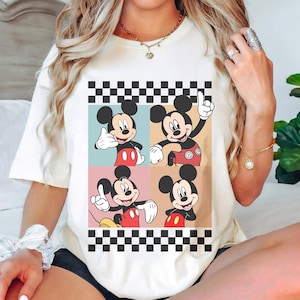 May include: Cream-colored t-shirt featuring a four-panel design of Mickey Mouse in various poses. Each panel has a different colored background, with a black and white checkered border at the top and bottom. Mickey is wearing his signature red shorts and white gloves.