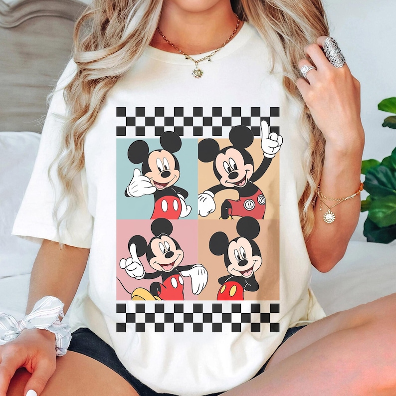 May include: Cream-colored t-shirt featuring a four-panel design of Mickey Mouse in various poses. Each panel has a different colored background, with a black and white checkered border at the top and bottom. Mickey is wearing his signature red shorts and white gloves.