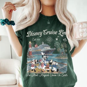 May include: Dark green t-shirt with a Disney Cruise Line graphic. The design shows a cruise ship, lighthouse, palm trees, fireworks, and Disney characters. Text on the shirt reads "Disney Cruise Line Est. 1998" and "The Most Magical Cruise On Earth."