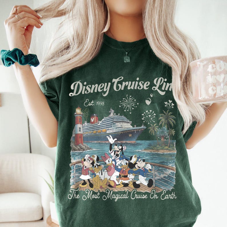 May include: Dark green t-shirt with a Disney Cruise Line graphic. The design shows a cruise ship, lighthouse, palm trees, fireworks, and Disney characters. Text on the shirt reads "Disney Cruise Line Est. 1998" and "The Most Magical Cruise On Earth."