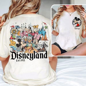 May include: Cream-coloured t-shirt featuring a Disneyland graphic on the back with Mickey Mouse, Donald Duck, and other Disney characters. The front of the shirt has a Mickey Mouse graphic with the word "Daddy".