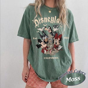 May include: A moss green short-sleeve t-shirt featuring a graphic of Mickey Mouse, Minnie Mouse, Goofy, Donald Duck, and a mummy character. The text reads Disneyland, Est 1955, and California. The design includes small bats and a distressed print style.