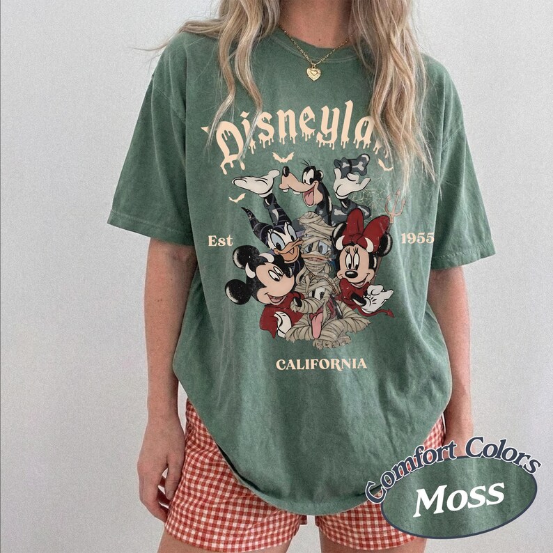 May include: A moss green short-sleeve t-shirt featuring a graphic of Mickey Mouse, Minnie Mouse, Goofy, Donald Duck, and a mummy character. The text reads Disneyland, Est 1955, and California. The design includes small bats and a distressed print style.
