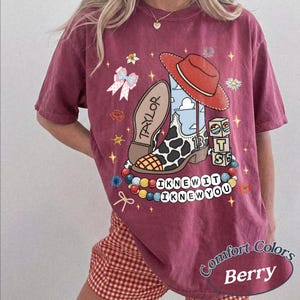 May include: A berry-coloured t-shirt featuring a graphic print of a cow-print cowboy boot, a red hat, a bow, and blocks with the letters T, S, and 13. The text "TAYLOR" and "I KNEW IT I KNEW YOU" are printed alongside colourful beads and small stars.