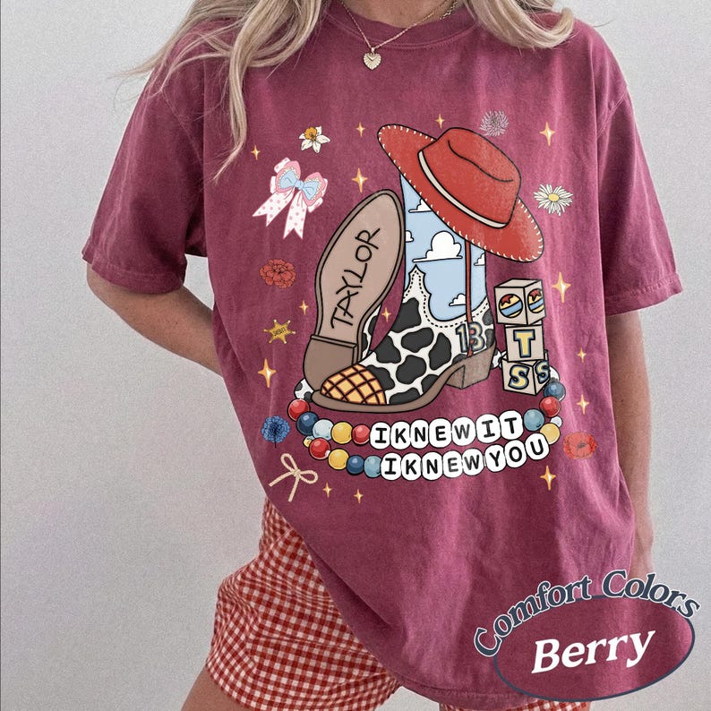 May include: A berry-coloured t-shirt featuring a graphic print of a cow-print cowboy boot, a red hat, a bow, and blocks with the letters T, S, and 13. The text "TAYLOR" and "I KNEW IT I KNEW YOU" are printed alongside colourful beads and small stars.