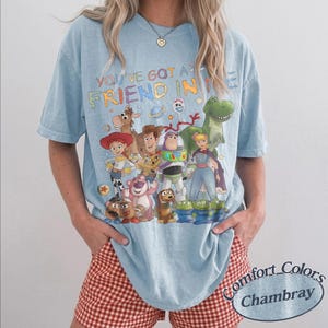 May include: A light blue short-sleeve t-shirt featuring a colorful graphic print of various Toy Story characters and the text "YOU'VE GOT A FRIEND IN ME" in a multi-colored font. The shirt has a crew neckline and a relaxed, loose-fitting silhouette.
