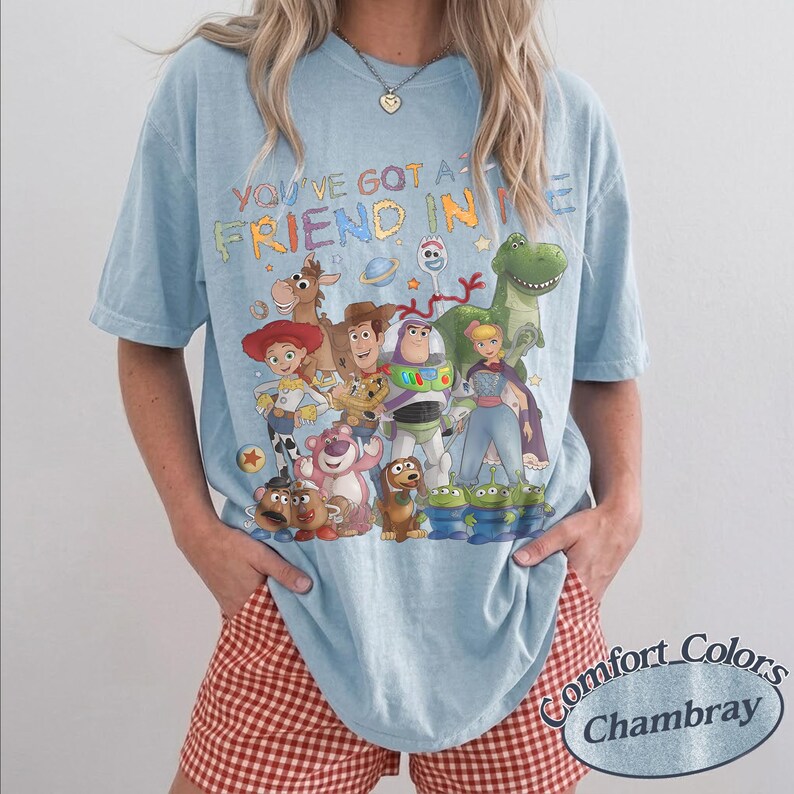 May include: A light blue short-sleeve t-shirt featuring a colorful graphic print of various Toy Story characters and the text "YOU'VE GOT A FRIEND IN ME" in a multi-colored font. The shirt has a crew neckline and a relaxed, loose-fitting silhouette.