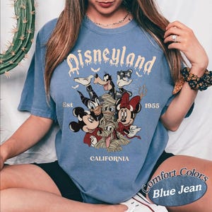 May include: A blue short-sleeve t-shirt featuring a graphic print of Disney characters including Mickey Mouse, Minnie Mouse, Donald Duck, and Goofy in costumes. The text reads Disneyland Est 1955 California with small bat silhouettes.