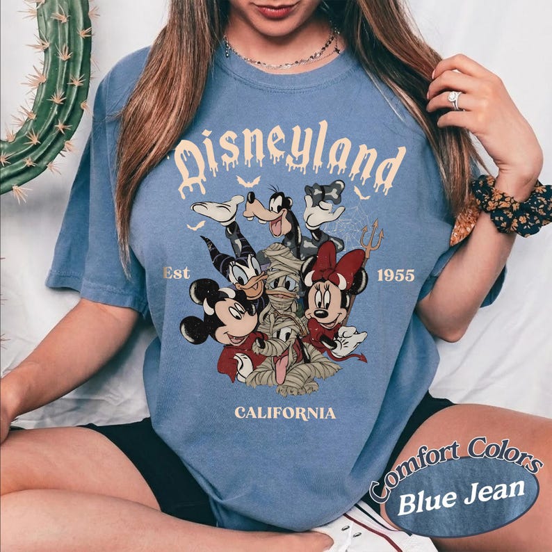 May include: A blue short-sleeve t-shirt featuring a graphic print of Disney characters including Mickey Mouse, Minnie Mouse, Donald Duck, and Goofy in costumes. The text reads Disneyland Est 1955 California with small bat silhouettes.