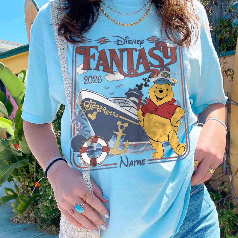 May include: Light blue t-shirt featuring a graphic of Winnie the Pooh in a sailor hat, a cruise ship, and the words "Disney Fantasy 2026". The shirt has a vintage travel poster design with a nautical theme.